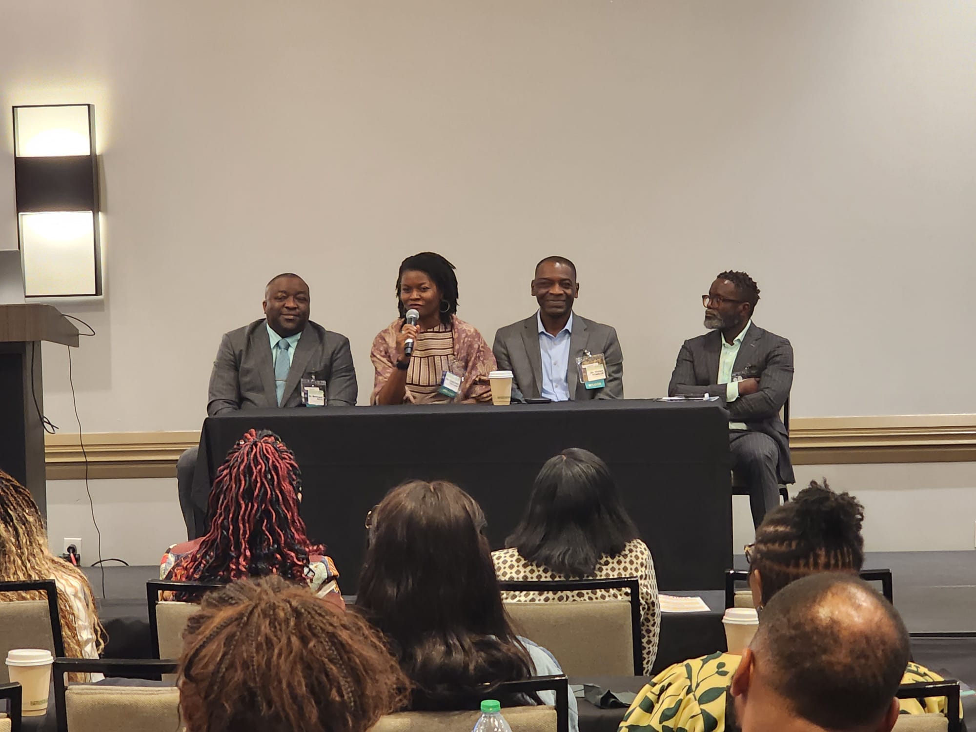 Panelists seated at a long table on a stage during a conference; a woman in the center speaks into a microphone while others listen, audience in foreground.
