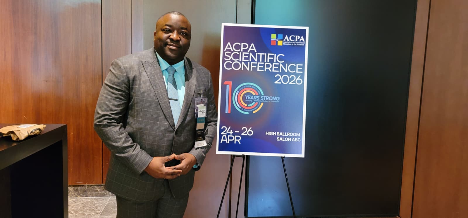 Man in a gray plaid suit posing next to a blue conference poster on a tripod stand for ACPA Scientific Conference 2026.