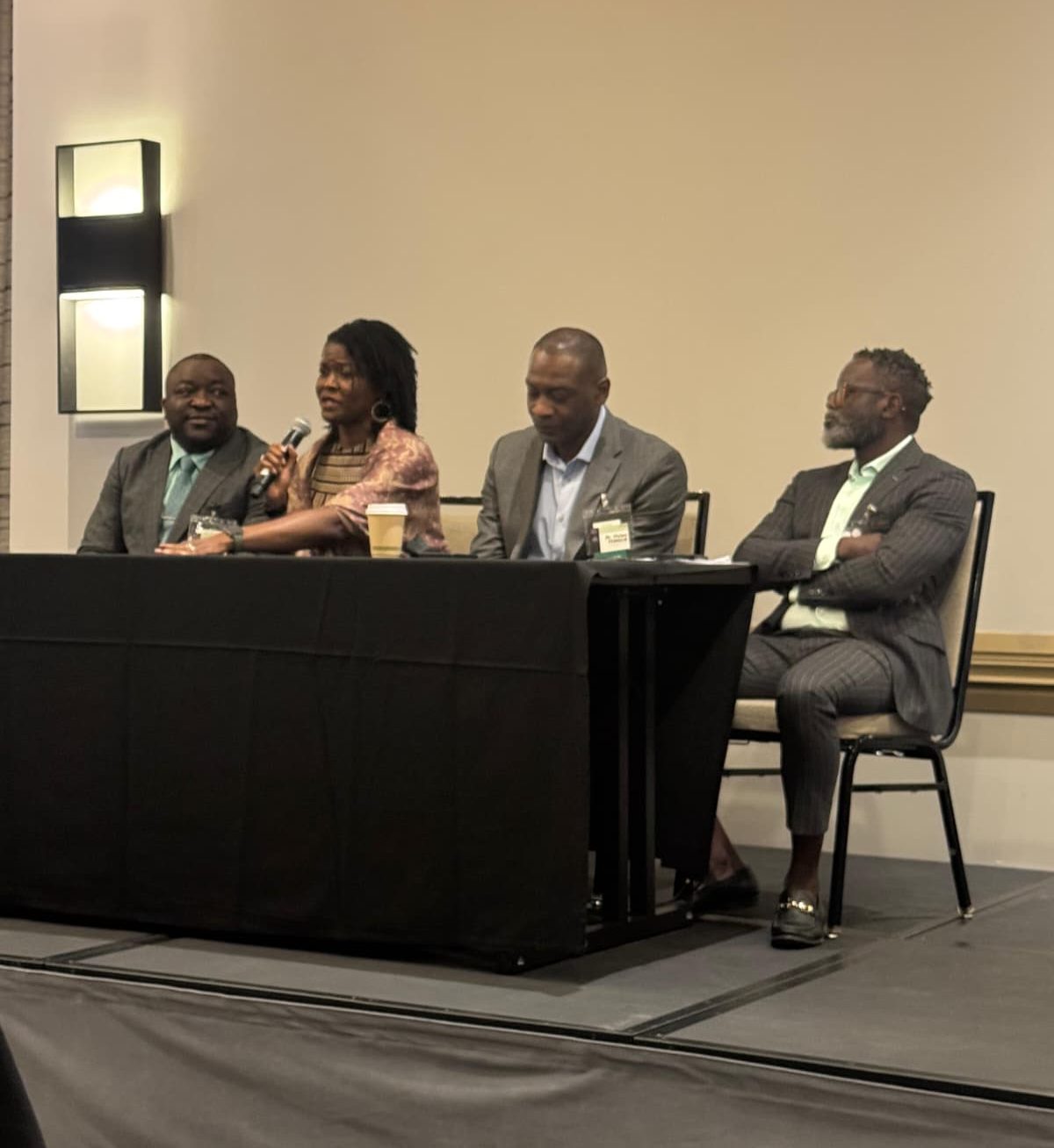 Panel discussion with four professionals seated behind a long black table; one woman speaks into a microphone while others listen, in a conference room setting with a cup of coffee on the table.