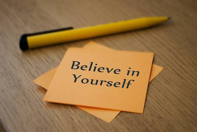 Sticky note with the message 'Believe in Yourself' on a wooden desk, a yellow pencil nearby — motivational reminder at a workspace.