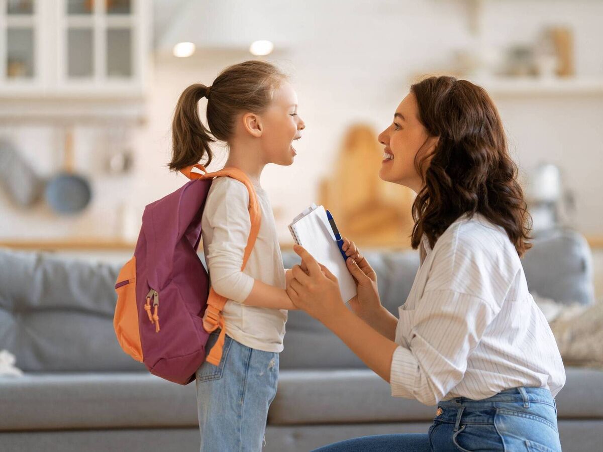 Happy family preparing for school - The Gatsby Showcase Foundation Mother kneels to speak with daughter who has a purple/orange backpack in a bright living room at home.