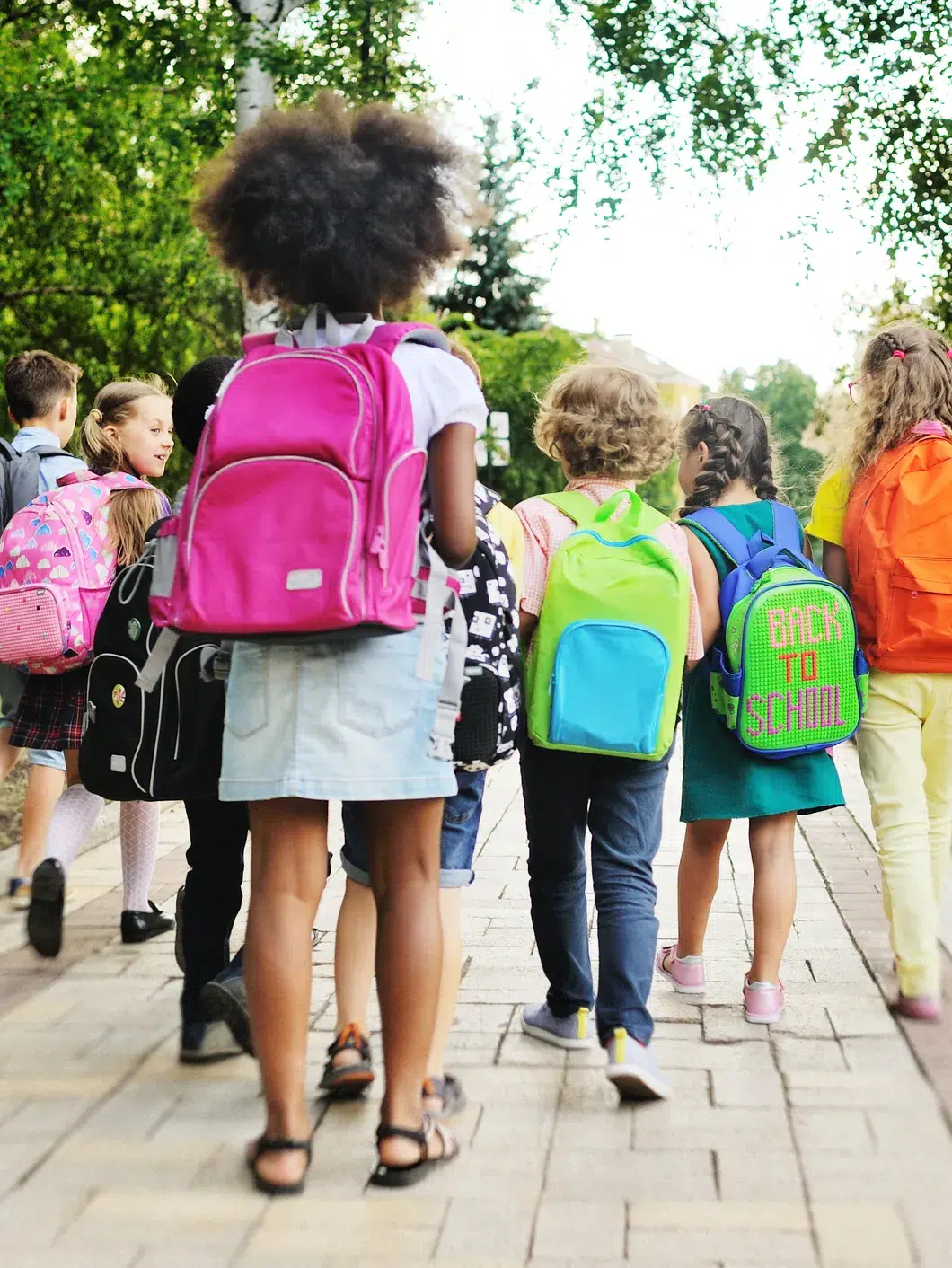 85321727007-getty-images-1327262834 - The Gatsby Showcase Foundation Group of children walking away on a sidewalk with colorful backpacks, heading to school in a sunny outdoor setting, trees in the background.