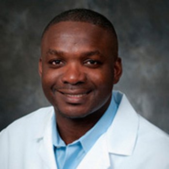 Portrait of a smiling Black male doctor in a white coat and light blue shirt against a neutral backdrop.