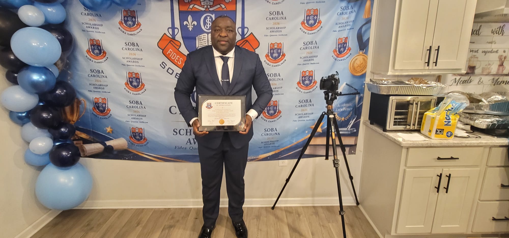 Man in a navy suit holding a framed certificate in front of a scholarship awards backdrop, with a blue balloon arch and a tripod camera nearby.