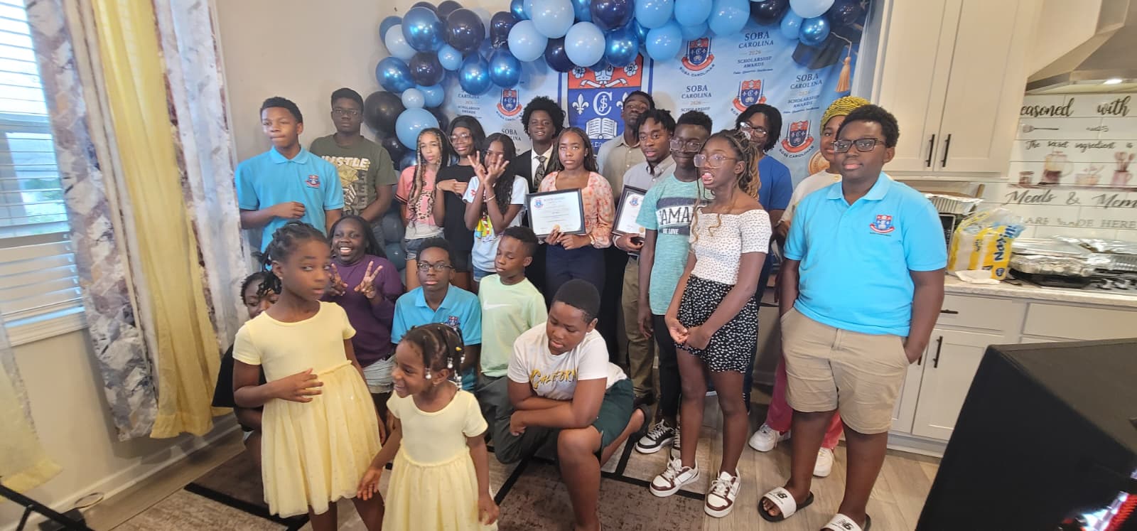 Group of diverse youths posing for a celebration photo with blue balloons and a banner in a home-style room; some hold certificates/screened awards.