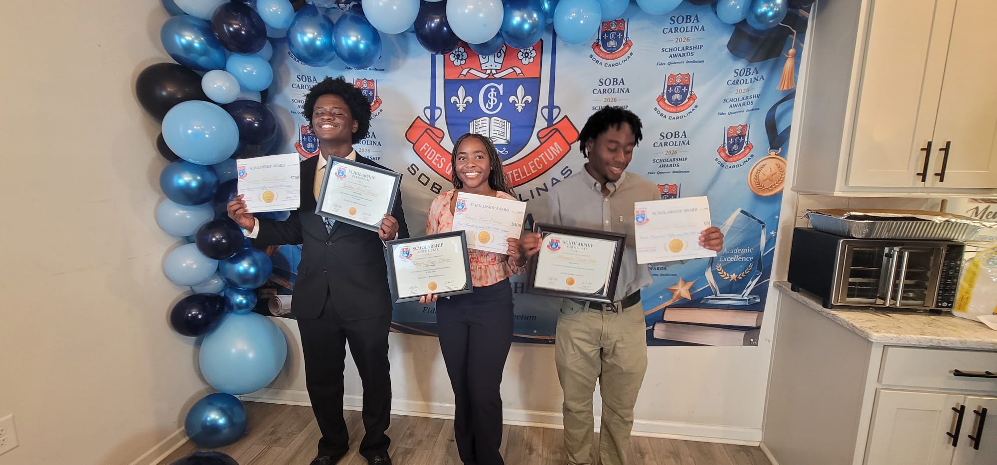 Three smiling students hold scholarship certificates in front of a blue balloon arch and school banner backdrop.