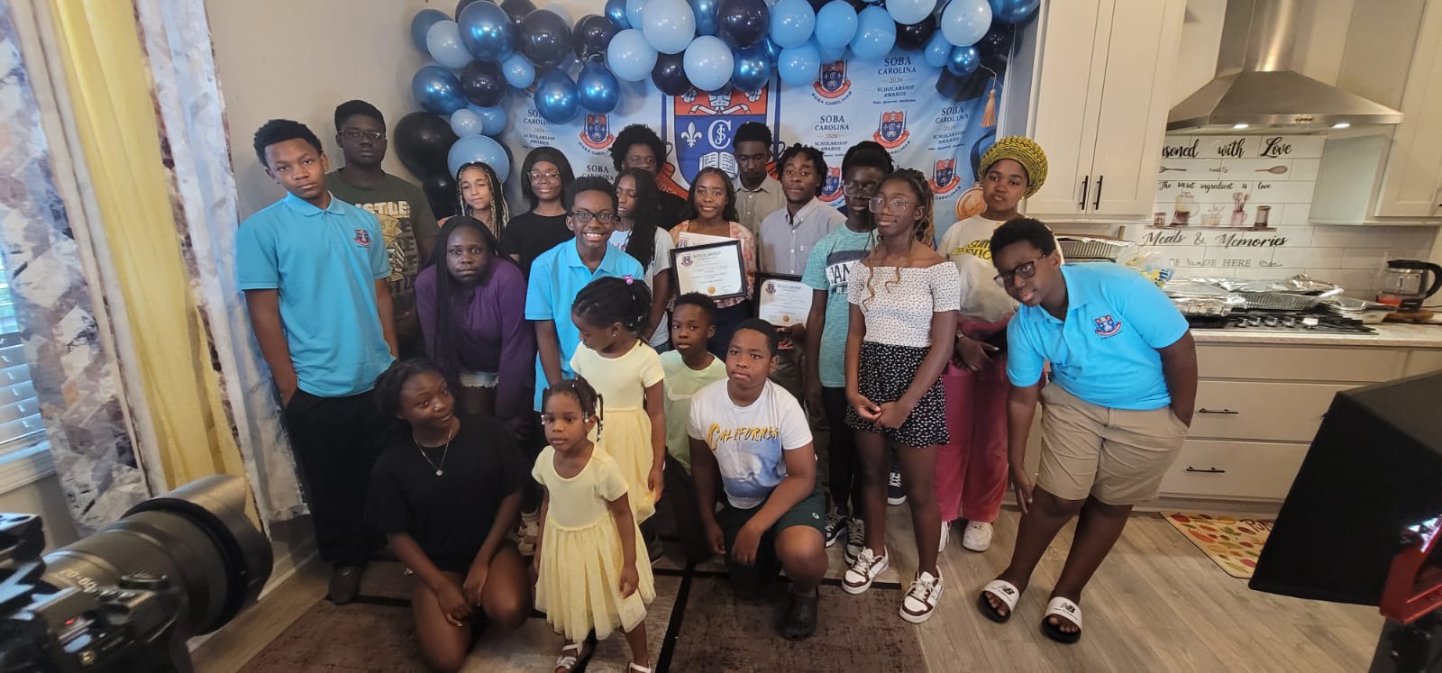 Group of teens and kids posing with certificates at an indoor celebration, blue balloons and a crest backdrop behind them.