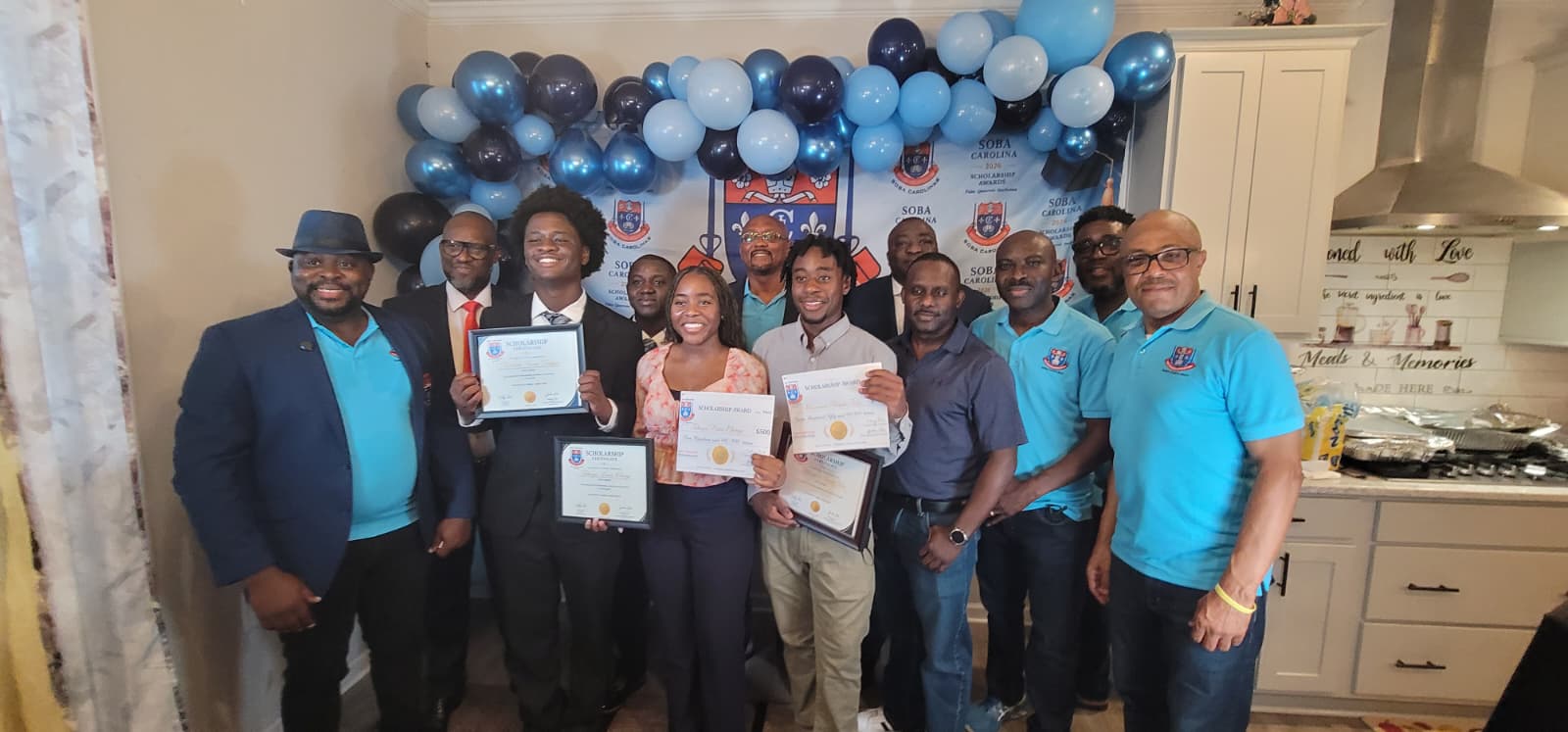Group of eleven adults posing with certificates at a celebration, blue balloons in the background and a kitchen setting nearby.