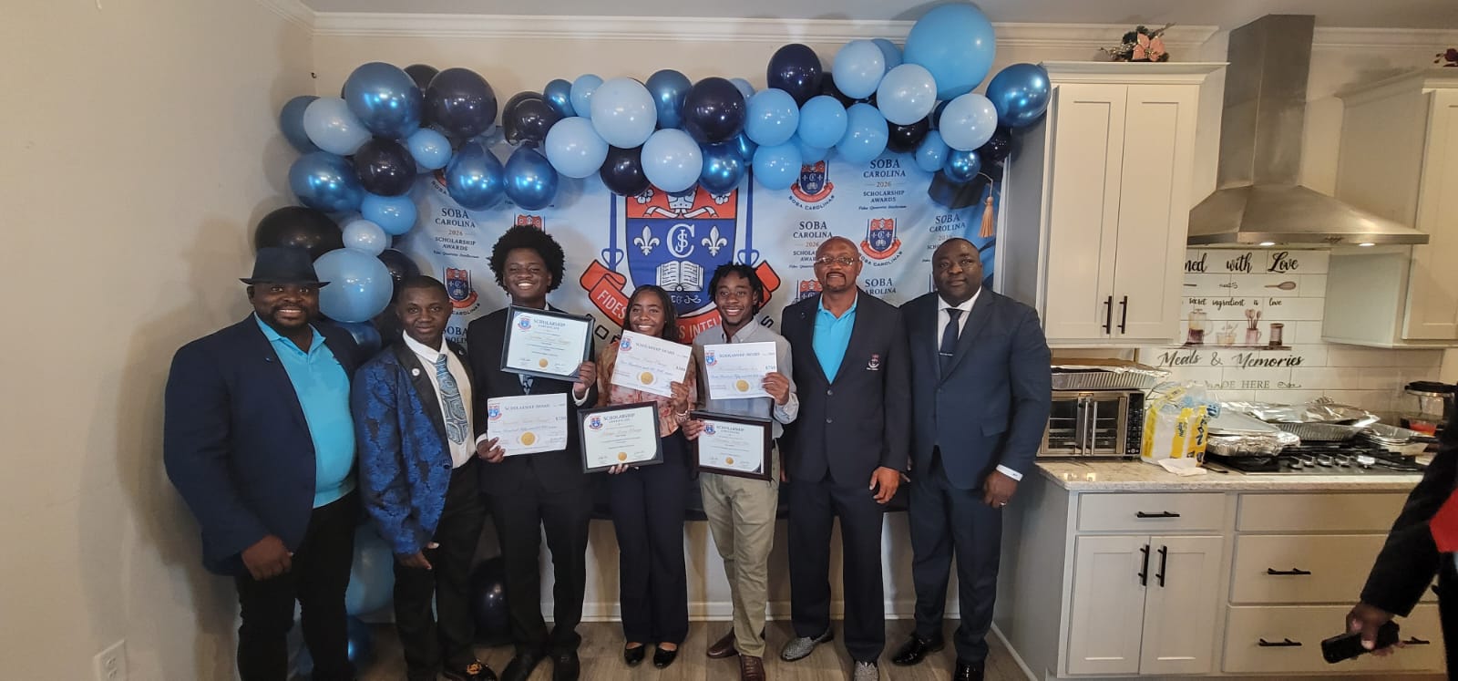Group of people posing with scholarship certificates at a ceremony, blue balloon arch and a banner in the background.