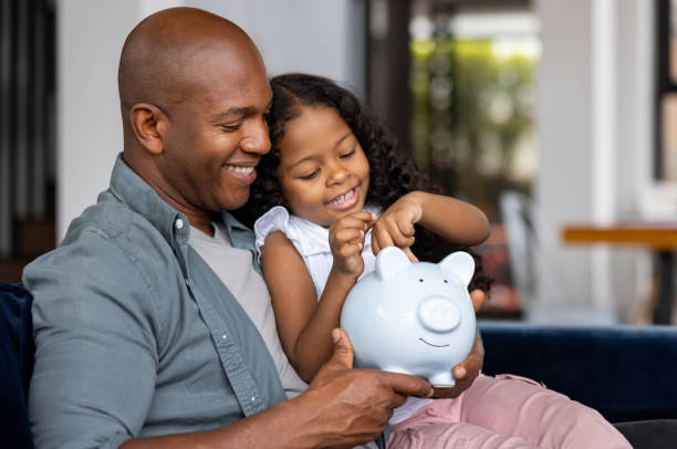 istockphoto-2065808904-612x612 - The Gatsby Showcase Foundation Father and daughter sit together, smiling as they place coins into a light blue piggy bank on the sofa indoors.