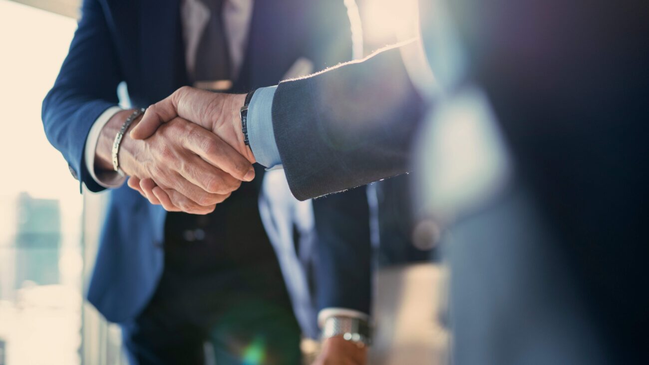 Business partnership meeting in office - The Gatsby Showcase Foundation Two businesspeople in suits shake hands in a bright office setting with a sunlit background.